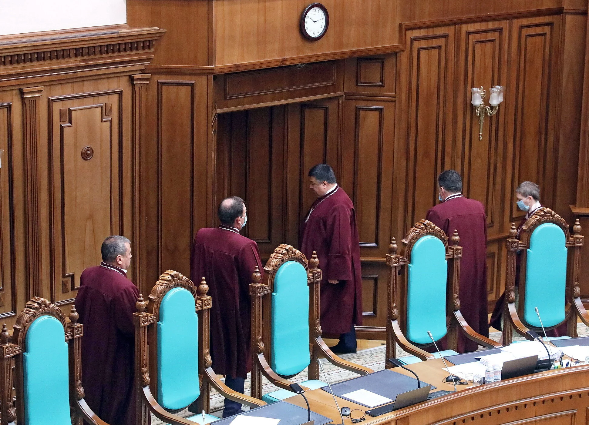 Судьи в бордовых мантиях стоят возле кресел в деревянном зале суда