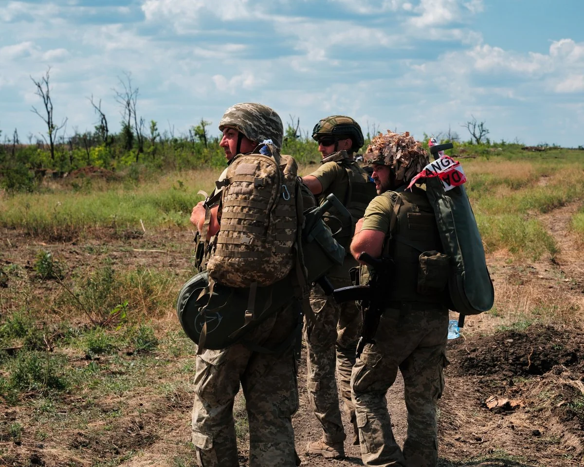 Троє військових у камуфляжі з рюкзаками стоять на ґрунтовій дорозі серед поля