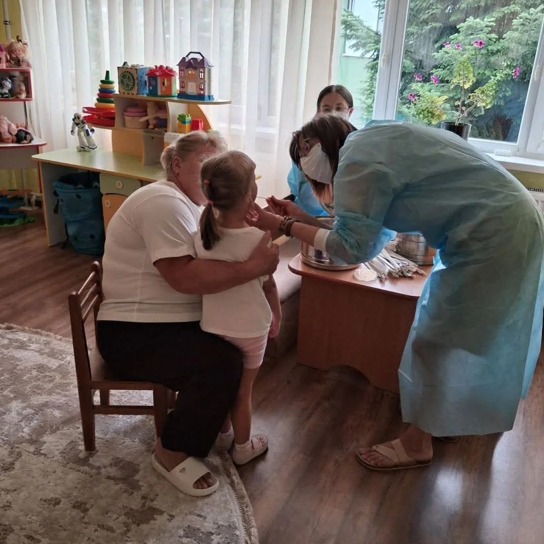 Жінка тримає дівчинку на колінах, медики в халатах обстежують її в дитсадку