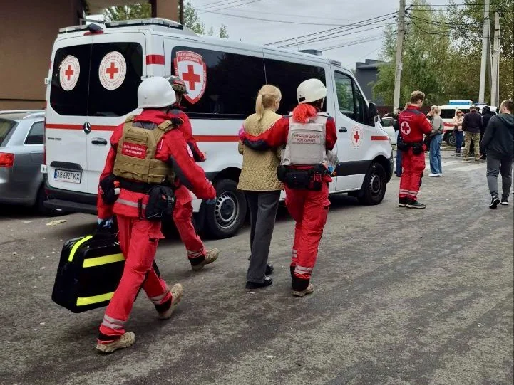 Рятувальники в червоній формі ведуть жінку до карети швидкої з червоним хрестом