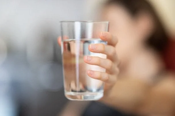 Рука тримає прозору склянку з водою на розмитому тлі людини