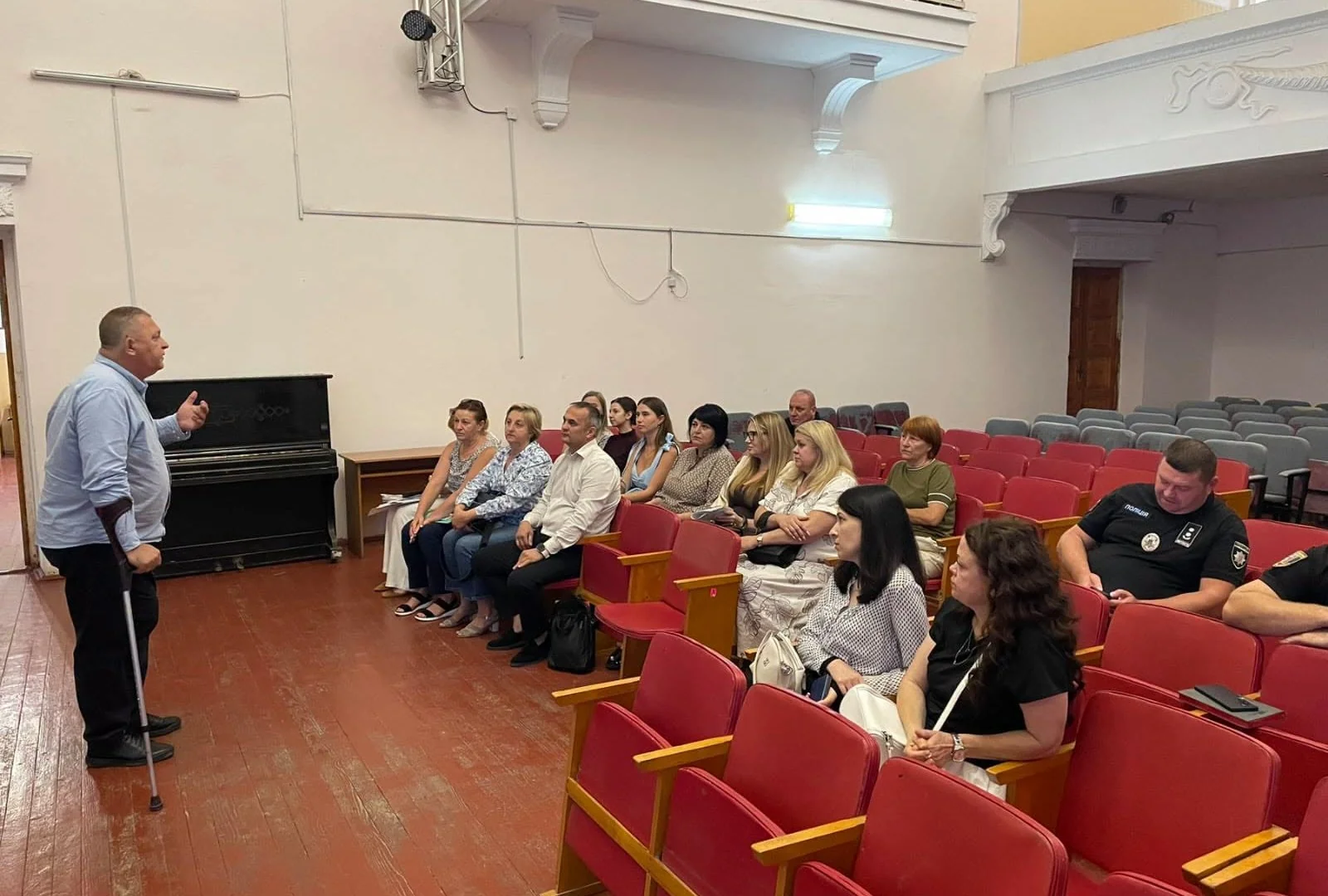 Чоловік з милицею виступає перед сидячою аудиторією в залі з червоними кріслами