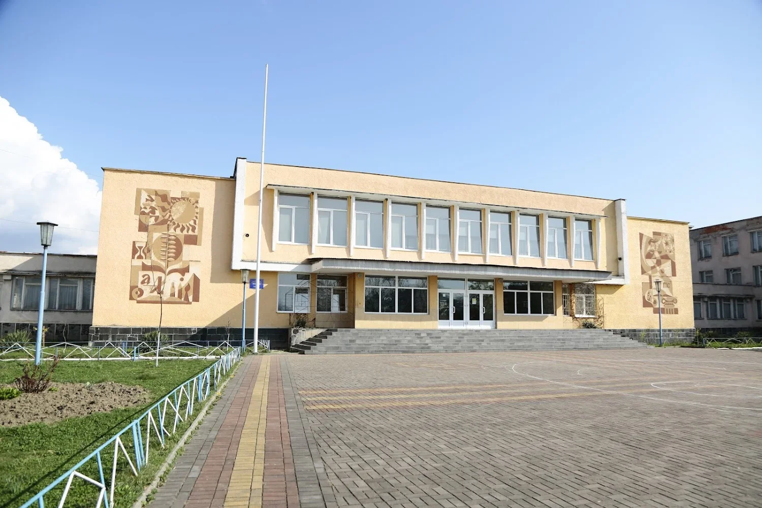 Жовта двоповерхова школа в Хусті з мозаїками на фасаді та брукованим подвір’ям