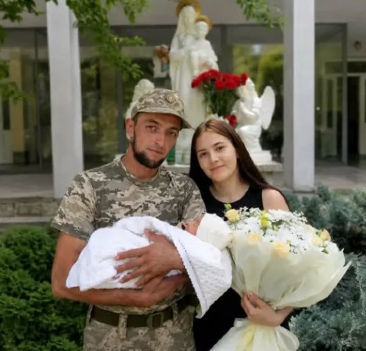 Військовий у камуфляжі тримає новонародженого, поруч дружина з букетом біля статуй лікарні