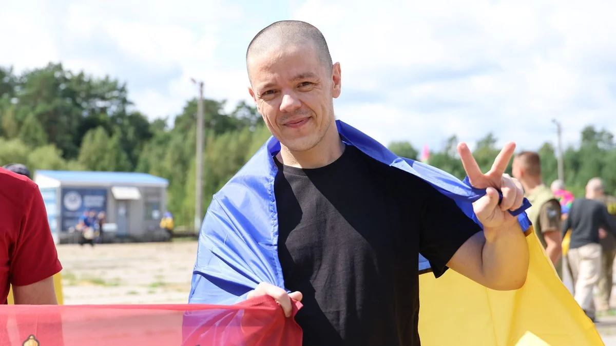 Усміхнений чоловік з виголеною головою в чорній футболці з українським прапором на плечах показує знак перемоги