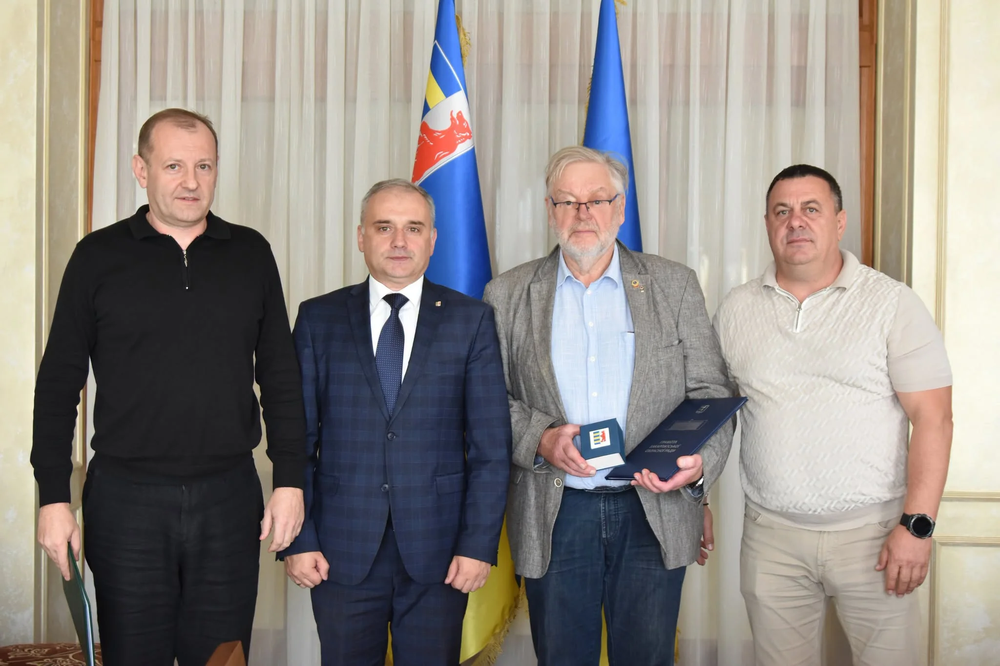 Чотири чоловіки на офіційній зустрічі з прапорами, один тримає грамоту і коробку