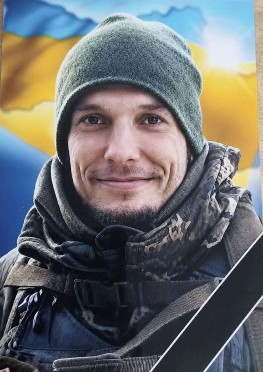 Усміхнений військовий у зеленій шапці та камуфляжі на тлі прапора України