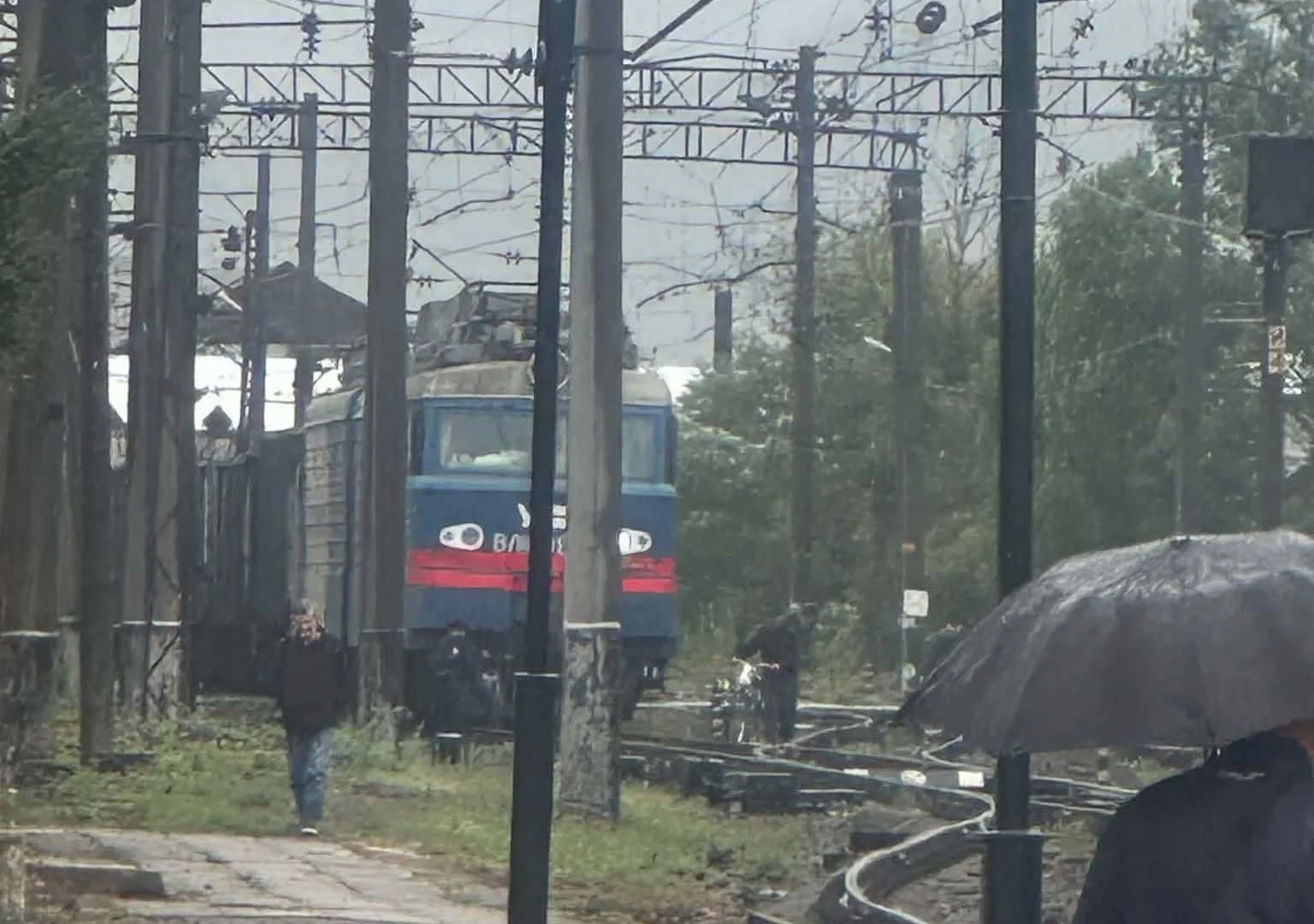 Синій електровоз на коліях під дощем біля пішоходів і парасольки