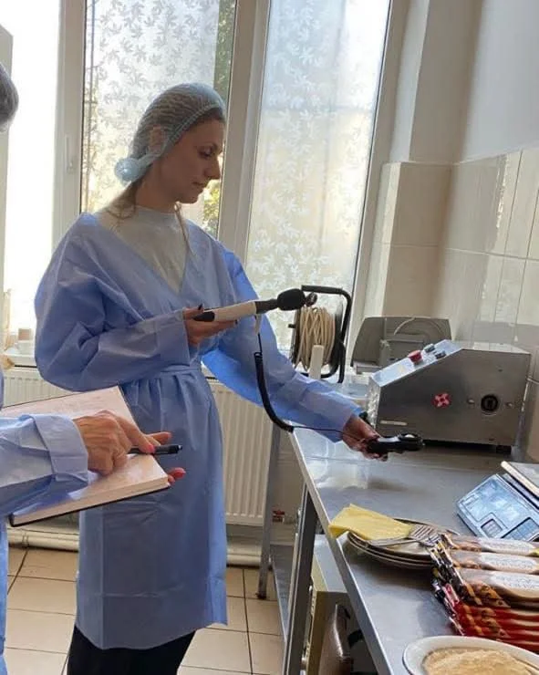 Жінка в халаті й сітці перевіряє кухню приладом, поруч ведуть записи