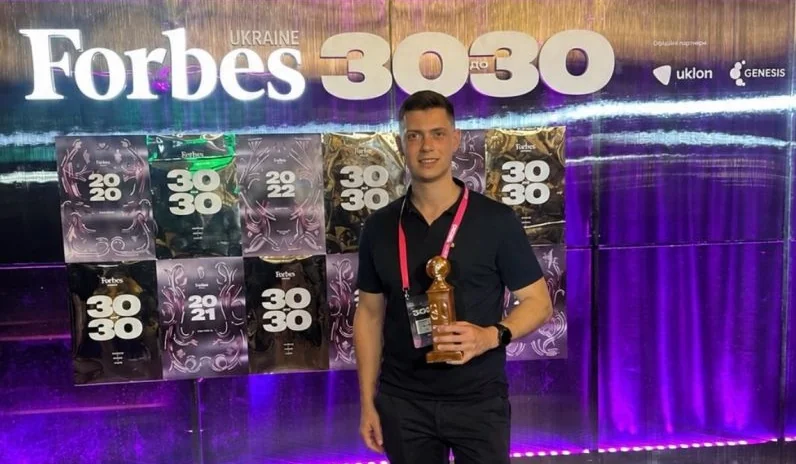 Усміхнений чоловік тримає кубок біля банера Forbes Ukraine 30 до 30